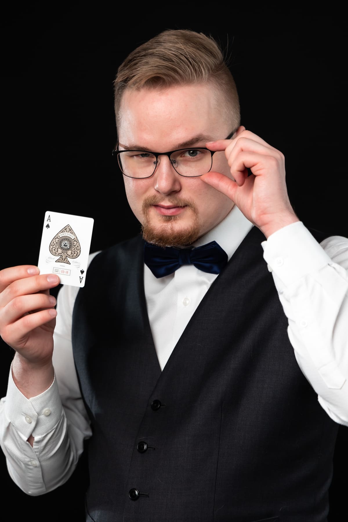 Magician Taavi Saarva holds up the ace of spades and looks at the camera over his glasses.