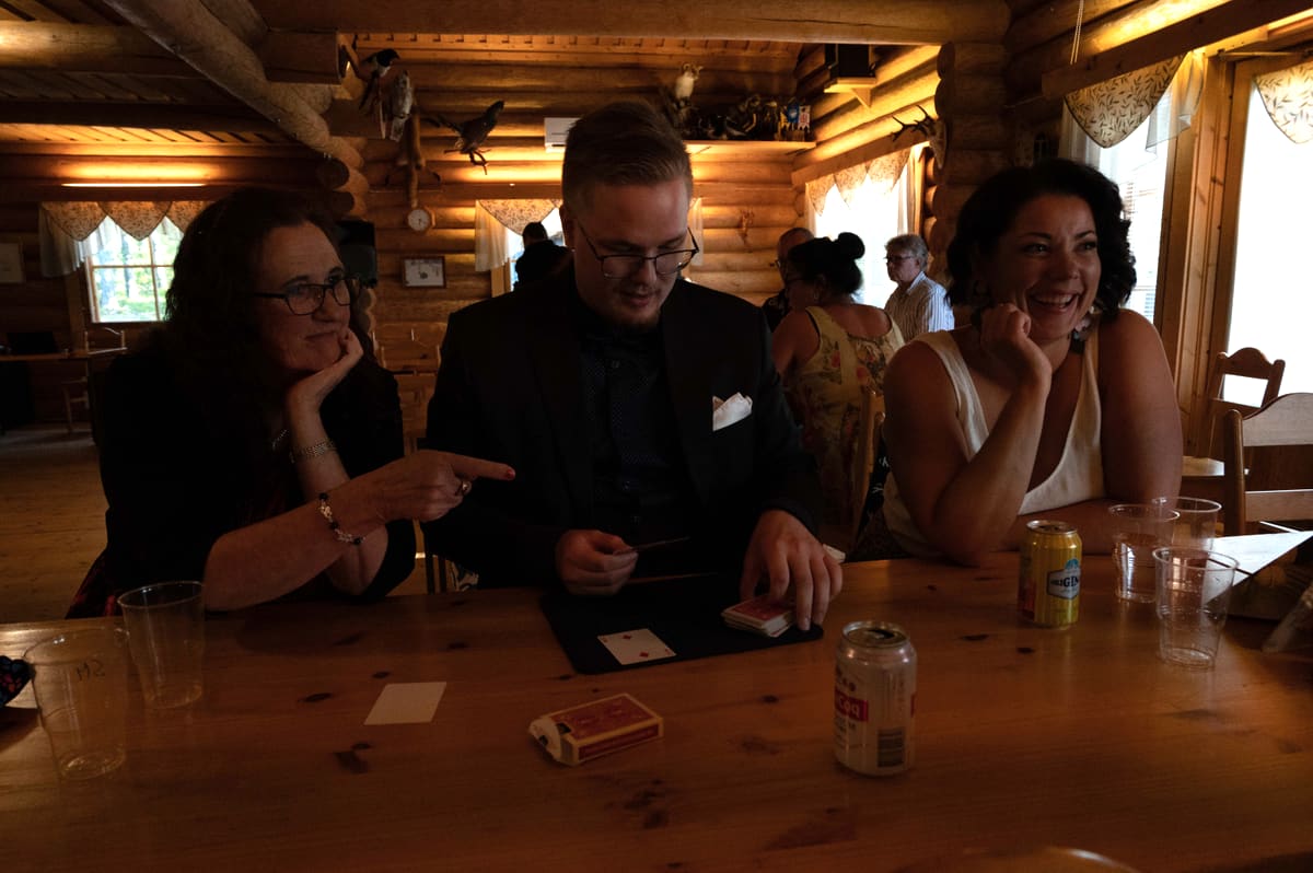 Taavi Saarva performs a card trick at a table while two guests laugh in amazement.