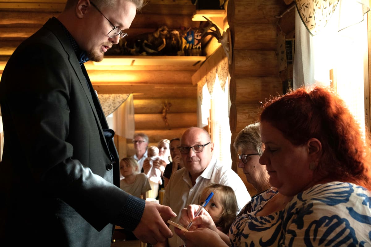 A guest signs a playing card held by the magician while a group of party guests looks on.